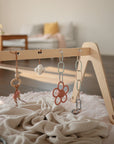 A mushie Daisy Rattle Teether hangs from a wooden baby gym with rings, set on a soft pink blanket in a cozy, sunlit living room with a couch in the background.
