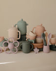 The Mushie Silicone Tea Play Set features pastel teapots, cups, saucers, spoons, plates, a flower-shaped teether, and a pacifier—perfect for play kitchen fun and imaginative pretend play on any beige surface.