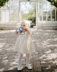 A young child in a white dress, bonnet, and tights stands on a sunlit stone path holding the mushie Daisy Rattle Teether, with greenery and a glasshouse in the background.