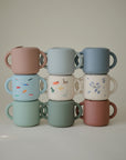 Nine mushie Snack Cups and mugs in pastel colors are stacked in rows. Some mugs have cute animal or plant prints; others are solid pink, green, blue, or cream—great for toddlers and pairing with the spill-proof silicone Snack Cup. The background is plain and light.