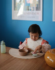 A young child plays with a mushie Suction Spinner Toy at a wooden table, wearing a bib. A bowl and water bottle sit nearby, while framed posters decorate the blue wall background.