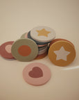 The mushie Foam Bath Puzzle features round, colorful tokens with printed hearts, stars, and circles, arranged on a neutral background for playful and educational bath time fun.