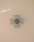 The mushie Suction Spinner Toy features a light gray, four-petaled flower design with a round center, made of silicone and mounted on a smooth beige surface, providing a calming sensory experience.