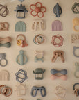 A selection of mushie Daisy Rattle Teethers and pastel silicone baby teething toys in shapes like rainbows, animals, flowers, and rings are neatly arranged in a grid pattern on a light background.