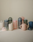 A collection of mushie Soft Spout Sippy Cups in spill-proof pastel shades—pink, blue, green, and white—is displayed on a neutral background with some matching lids beside each cup.