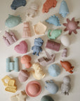 Lifestyle image of a collection of colorful silicone bath play set toys on a light background. The mix includes playful shapes from the Sealife, Pirate, Princess, Vehicles, Space and Dinosaur sets.