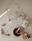 The mushie Silicone Baby Bib, shown with a handled cup, bowl, spoon, and beaded pacifier clips on a cream mat, is easy to clean and has an adjustable neck strap plus charming animal and fairy designs in muted colors.