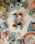 Mushie Pour and Play Bath Set includes pastel cups for sensory bath play on a light surface.
