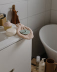 A modern bathroom features a white sink cabinet, bath products, a cupcake-shaped soap, heart-shaped soap, and the Mushie Organic Cotton Bath Mitt—ideal for sensitive skin—beside a towel and a freestanding white bathtub in the background.