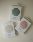 Three boxes of mushie Muslin Nursing Cover in green, beige, and pink rest on a light beige surface. Each box features a circular window showing the soft fabric inside.