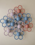 A set of mushie Daisy Rattle Teethers, featuring pastel blue, pink, and purple flower shapes, closely arranged on a light beige surface—ideal as a gentle baby teething toy or silicone rattle teether.