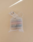 A mesh drawstring bag with mushie Foam Bath Puzzle toys sits on a beige surface. An attached tag features text and colored circles, highlighting the educational benefits of these playful items.