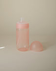 The mushie Soft Spout Sippy Cup, featuring a translucent pink body with a matching silicone nipple and separate rounded cap, rests on a plain light surface against a neutral background.