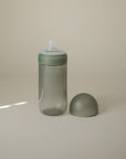 The mushie Soft Spout Sippy Cup, featuring a translucent gray body, green cap, and clear silicone nipple, stands upright on a light surface with its matching rounded cap beside it. The background is softly lit and neutral.