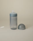 The mushie Soft Spout Sippy Cup, a translucent gray toddler cup with a blue-gray lid and silicone spout, stands upright on a beige surface with its rounded cap beside it—designed for easy, comfortable sipping.