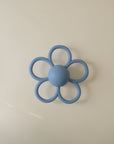 The mushie Daisy Rattle Teether features a blue, flower-shaped design with a textured surface and round center, shown on a plain light background.