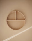 The mushie Silicone Suction Plate, round and beige with three compartments, rests on a smooth light beige surface highlighted by a diagonal streak of light.