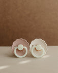 Two FRIGG Daisy Night Silicone Pacifiers, one pale pink and one white, with scalloped edges and round handles, rest side by side on a soft beige surface against a muted brown background.