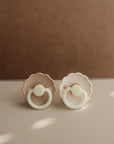 The FRIGG Daisy Night Silicone Pacifier 2-Pack features two pastel pacifiers with scalloped edges and round handles, displayed on a light surface against a soft brown background.