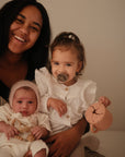 A smiling woman holds a baby in a knit bonnet and beaded pacifier clip, as a toddler with a FRIGG Natural Rubber Pacifier from the FRIGG 4-Pack sits on her lap, wearing a ruffled white shirt and holding a pink sippy cup.