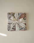A folded Muslin Cotton Washcloth by mushie, featuring a floral pattern and tied with a white ribbon printed with the brand name, rests on a light beige surface—ideal as a soft, organic cotton baby washcloth.