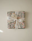 The Muslin Cotton Washcloth by mushie features a soft floral pattern and is folded neatly, tied with a white ribbon printed with the mushie logo, displayed against a neutral background.