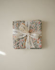 A folded Muslin Cotton Washcloth by mushie with a floral pattern and tied with a white mushie ribbon rests on a plain, light-colored surface, casting a soft shadow in the corner.