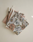 Four mushie Muslin Cotton Washcloths—two with floral patterns in muted pink, yellow, and green, and two in solid pastels—are stacked neatly with white loops on a light surface.