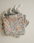 A stack of mushie Muslin Cotton Washcloths in soft, muted floral patterns, each with a small white hanging loop, arranged on a light surface—combining style and gentle organic cotton comfort.