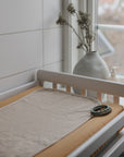 A neatly arranged Mushie Changing Pad Liner sits on a beige mat with a teething ring. In the background, natural light streams in, highlighting a vase of dried flowers and a bottle on the windowsill.
