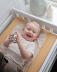 A smiling toddler lies on a yellow-covered changing pad, holding a toy. The table by the window features Mushie Changing Pad Liners, with lotion and a vase placed on the windowsill.