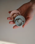 A hand holds two FRIGG Rope Natural Rubber Pacifiers by Mushie—one pastel pink, one pastel blue—both with textured edges and natural rubber latex nipples; a perfect baby essential 2-pack.