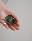 A hand holds a FRIGG Rope Silicone Pacifier from the FRIGG Rope Silicone Pacifier 2-Pack, featuring medical grade green silicone with a circular, textured rim and two holes near the center, against a neutral background.