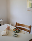 A wooden high chair is placed at a white table set for baby feeding with a mushie Silicone Suction Plate filled with cut veggies and fruit, silicone bib, pink sippy cup, and flower-shaped mat. A lamp and framed art accent the minimalist room.
