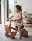 Smiling toddler rides a pink mushie Mini Toddler Bike indoors by a bright window.