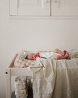 A newborn in a white onesie and gray hat rests on the mushie Extra Soft Muslin Changing Pad Cover, atop a checkered pillow and striped blanket. Below the table, a stack of diapers completes the serene, softly lit scene.