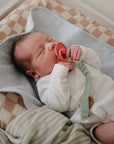 A newborn sleeps peacefully on neutral-toned bedding, holding a pink pacifier while lying on Mushie Changing Pad Liners, dressed in a white organic cotton onesie atop a checkered blanket.