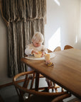 A young child with light blonde hair sits at a wooden table, eating from the mushie Round Dinnerware Bowl. Warm sunlight streams through a window, casting shadows. A textured woven wall hanging decorates the space behind the child.