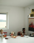 Four young children gather around a white table in a bright, modern dining room, eating from mushie Square Dinnerware Plates while drawing. The room features minimal decor, a scenic window view, and a brass ceiling lamp overhead.
