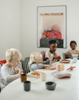 Four children sit at a white table eating watermelon, using mushie Round Dinnerware Plates and colorful cups. Sunlight fills the room, and a vibrant painting decorates the wall behind them.