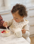 Toddler eats fruit with Rylee + Cru Feeding Kit Heart Ditsy, wearing a silicone bib at counter.