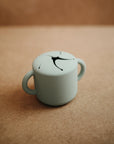 The mushie Snack Cup is a small, light green silicone cup with two handles and a spill-proof petal lid, ideal for toddlers. It sits on a brown surface.