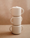 Three white ceramic mugs with handles are stacked vertically against a beige background, perfectly paired with the mushie Snack Cup—a spill-proof silicone essential for toddlers and busy families.