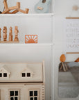 A neatly organized children's room shelf featuring mushie's Sun Teether, wooden toys, a small dollhouse, a blue ukulele, an alphabet wall hanging, and a mirror with orange animal ears.