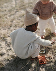 Two young children in knit sweaters and hats play outdoors on sandy ground with toys, including the mushie Sun Teether, surrounded by sparse grass.