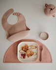 The mushie Snack Cup is a muted pink toddler feeding set, including a silicone bib, spill-proof cup, and bowl on a placemat—plus a matching snack cup for treats. The bowl holds heart-shaped pancakes, banana slices, raspberries, and syrup.