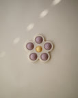 The mushie Flower Press Toy features five pinkish-purple petals and a yellow center, designed to support sensory exploration and fine motor skills, shown on a plain light background with spots of sunlight.