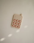 The mushie Phone Press Toy, in beige and pink with rounded buttons and a calculator-like design, rests on a light surface—ideal for sensory play and developing fine motor skills.