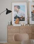 A modern wooden sideboard with framed abstract art, a black floor lamp, a tan vase, books, decorative objects—including a white speaker, candlesticks, and the Mushie Silicone Tea Play Set—stands against a neutral wall.