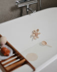 A white tub with a small amount of water features the mushie Water Wheel Bath Toy, shaped like a beige flower, and a matching scoop. A wooden tray holds pastel bath items and a candle. The faucet is set in a gray tiled wall.